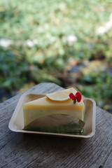 A slice of lemon mousse cake with a glossy finish, garnished with a lemon wedge and red flower, served on a paper tray atop a rustic wooden table. Natural green background, bokeh style.