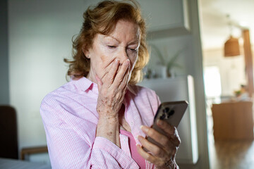 Shocked senior woman reading unexpected message on smartphone at home