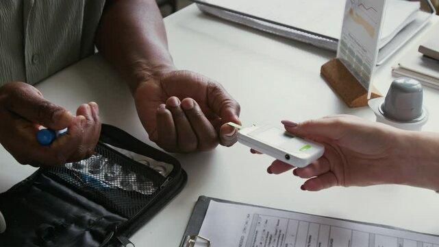 Close-up of mature African American male patient pricking his finger with pen while clinician checking blood sugar level with glucometer by workplace in clinics