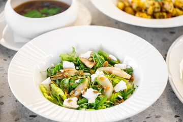 Fresh salad with mixed greens, pear slices, feta cheese, and seeds in a white bowl, accompanied by a bowl of soup and roasted vegetables. Healthy and nutritious meal.