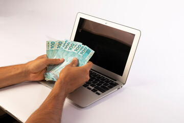 hands of a man holding Brazilian banknotes in front of a computer white background