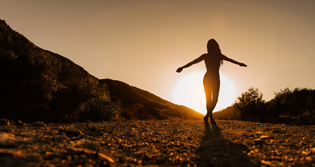 Silhouette of a young woman balancing with open arms on mountain path at sunset.
