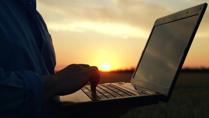 Laptop in hands of businessman outdoors, close-up. Businessman farmer with laptop working on field, estimating grain harvest. Farmer man working on computer, sunset. Agricultural business concept