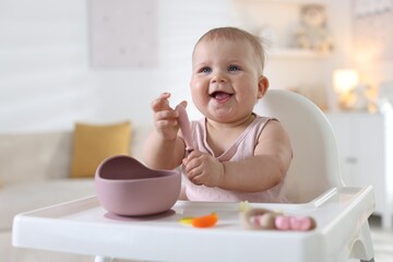Cute little baby eating with spoon on feeding chair at home. Space for text