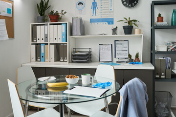 Office break room featuring table with snacks, coffee cup, and hygiene supplies amid organized shelves and health poster promoting wellness in workplace setting