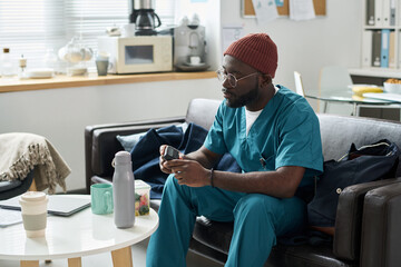 Wearing teal scrubs and a red beanie, an individual using a smartphone in a modern office with various devices and files organized around them creating a productive atmosphere