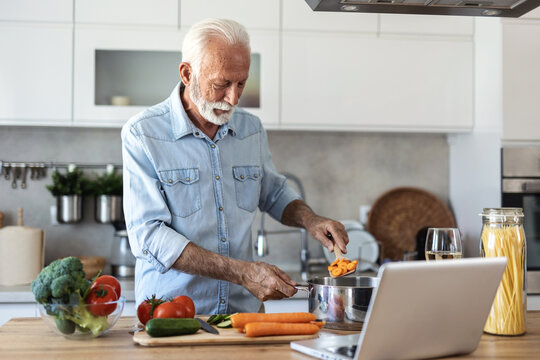 Happy senior man cooking healthy food with recipe. Senior man makes breakfast by looking at how to make it with his laptop computer with a happy face. At her own house.
