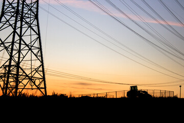High-voltage power lines stretching across a vivid sunset sky, with the silhouette of a tractor...