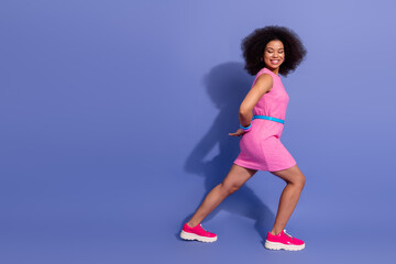 Energetic woman in a pink dress walking confidently against a vibrant purple backdrop in a stylish,...
