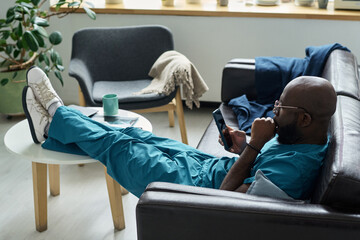 Man lounging on modern sofa while looking at smartphone, enjoying a moment of relaxation. Living room decor includes houseplants and cozy furniture