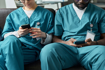 Two medical professionals in blue scrubs sitting on a couch using smartphones. One person wearing...