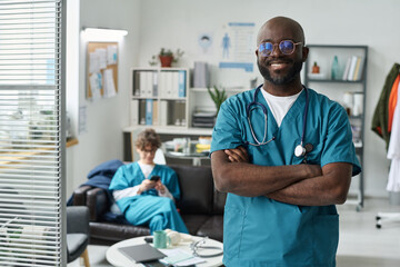 African American doctor standing with arms crossed, smiling confidently in modern medical office, while a nurse sits on couch in background, busy with paperwork
