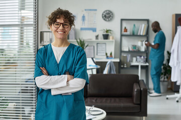 Healthcare worker standing with crossed arms in a medical office, smiling. Background includes a coworker working on documents and medical equipment, creating a professional environment