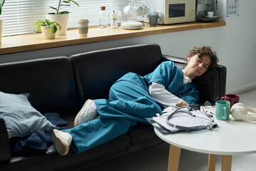 Nursing professional in scrubs taking short rest on sofa in hospital break room Featuring medical equipment and personal items on table nearby
