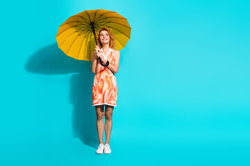 Smiling young woman in a vibrant summer dress holding a yellow umbrella against a turquoise...