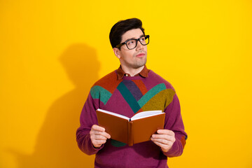 Young man in glasses holding a book and pondering against a vibrant yellow background, showcasing style, intellect, and retro charm.