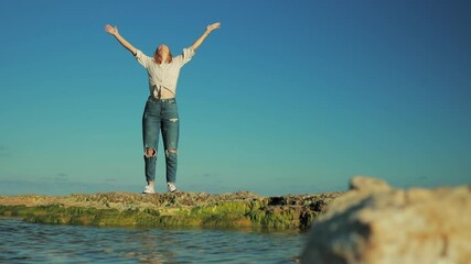 Woman traveler stand on rocky shoreline, spreading arms wide and absorbing expansive ocean vista with sense of liberation and inner peace beneath vibrant blue sky. Lifestyle, vacation, tourism, nature