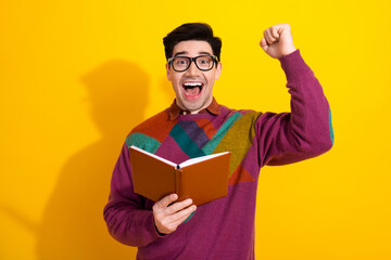 Excited young man holding a book with joy against a vibrant yellow background, wearing colorful...