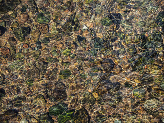 Numerous smooth, rounded pebbles of varying sizes and colors, submerged in clear, shallow water. View of the ripples of water flowing over colorful pebbles in a shallow river bed