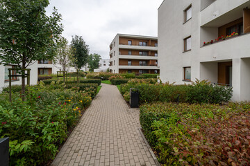 Landscaped pathways wind through this modern residential complex courtyard. Dense shrubs, ornamental grasses, and trees line paved walkways between apartment buildings, displaying seasonal color.