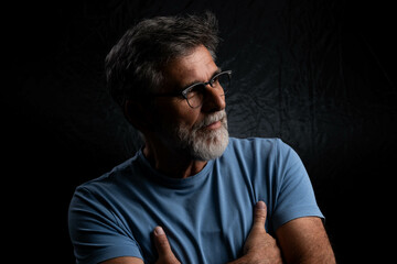 Handsome old-fashioned hipster in a t-shirt posing with crossed arms. Muscular mature man with stern expression standing confidently against dark background.