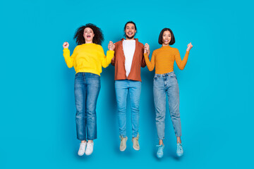Fototapeta premium Three young friends in casual clothing jumping joyfully against blue background, celebrating friendship and fun.
