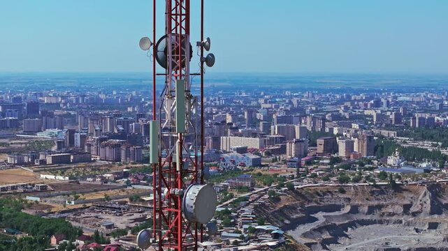 Aerial close-up of a large microwave antennas mounted on a red and white telecommunication tower. The structure stands against the cityscape of Bishkek, showcasing urban infrastructure.