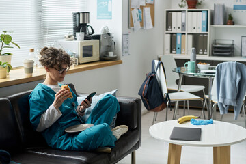 Nurse relaxing in break room, holding phone and snack. Tables filled with food and drinks, office supplies organized on shelves. Natural light filtering through window enhancing ambiance