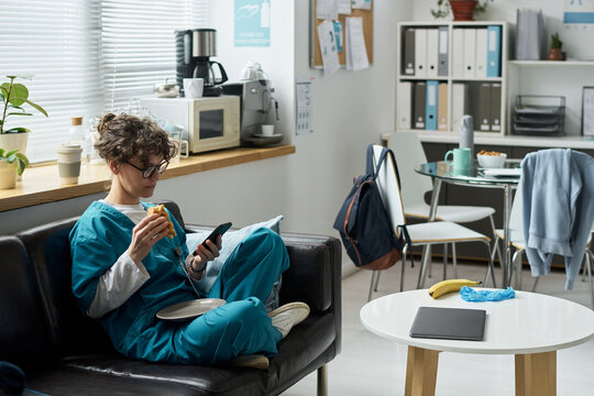 Nurse relaxing in break room, holding phone and snack. Tables filled with food and drinks, office supplies organized on shelves. Natural light filtering through window enhancing ambiance - Powered by Adobe