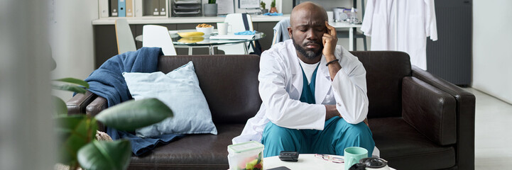 Black person dressed in medical attire sitting on couch while holding head, appearing tired and...