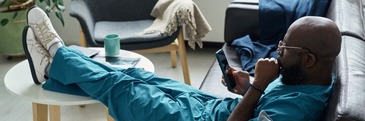 Healthcare worker wearing scrubs reclining on couch and engaging with tablet device in a modern, relaxed setting showing a moment of leisure and technology integration