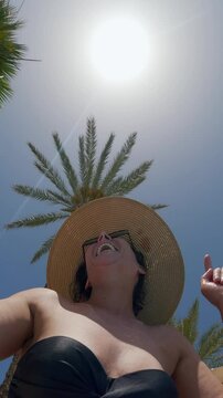 Positive smiling fashionable woman wearing a hat and sunglasses enjoys dancing outdoors amidst tropical palm trees, vertical video