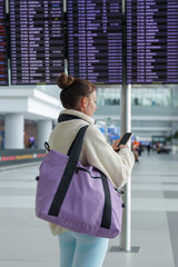 Obraz premium A young traveler stands in a modern airport terminal, checking flight information on a mobile device while holding a large purple bag. The digital display shows various flight statuses.