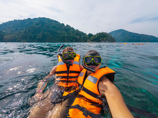 Two friends enjoy snorkeling in Thailand, wearing bright orange life vests while exploring vibrant...