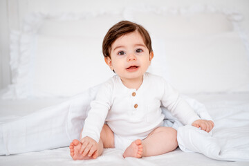 A baby under a blanket on a white bed at home, a place for text, a cute smiling little child getting ready for bed