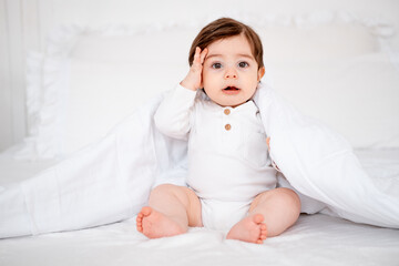 A baby under a blanket on a white bed at home, a place for text, a cute smiling little child getting ready for bed