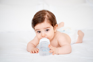 baby with a bottle of milk or water on a white bed at home, baby food feeding concept, place for text, cute smiling little baby