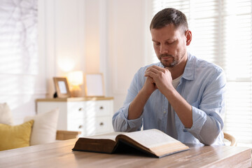 Man with Bible praying at table indoors. Space for text
