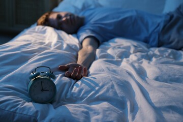 Frustrated man suffering from insomnia on bed indoors, focus on alarm clock