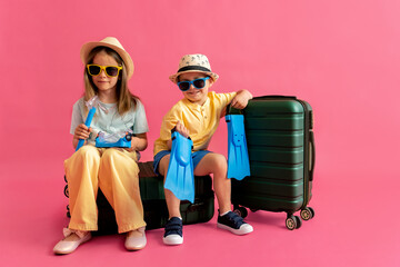Boy with flippers and girl with swimming mask sitting on green suitcase on pink background. Children playing going on vacation to the sea. Concept of summer vacation, travel.