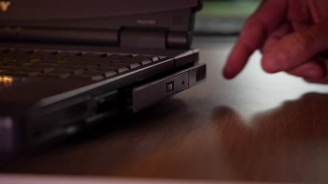 A close-up shot captures the precise and functional mood as a hand carefully inserts a CD into an older model laptop's optical drive, in a softly lit setting