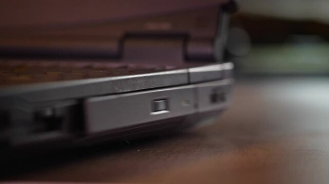 A close-up shot captures the precise and functional mood as a hand carefully inserts a CD into an older model laptop's optical drive, in a softly lit setting