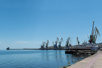 View of seaport in city against cloudy sky. Port cranes are located along the coastline. In foreground is emerald water of Black Sea. Feodosia port