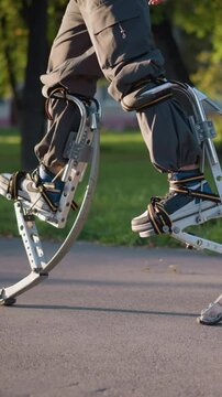 Legs of man using spring stilts while walking on paved path in sunny park, wearing dark gray pants. Low camera angle focuses on dynamic movement, highlighting stilts and vibrant outdoor scenery