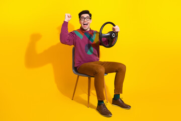 Young man with steering wheel on yellow background expressing excitement and joy, wearing casual...