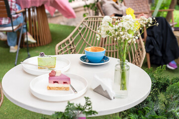 A cozy outdoor caf? table featuring two colorful desserts, a cup of coffee, and a vase of flowers, set against a vibrant garden background, offering a delightful and relaxing atmosphere.