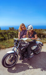 Obraz premium Two women, one of them sitting on her chopped motorcycle, consulting a map on a picturesque coastal road with the sea in the background.