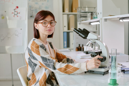 Portrait of student wearing glasses, working with microscope in science laboratory filled with equipment and bookshelves. Background including whiteboard with chemical formulas and organized shelves