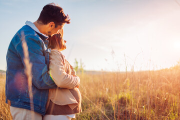 Portrait of pregnant family couple outdoors. Future dad hugging his wife from behind. Woman showing her baby bump
