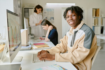 Smiling African American student working on computer in modern lab with classmates in background engaging in various tasks. Visible computer screen showing colorful graph